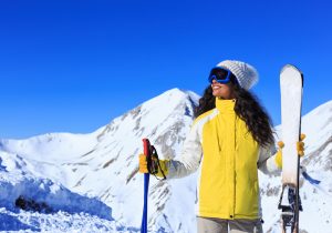 Woman with yellow jacket smiling and enjoyng her winter vacation, holding ski equipment on a mountain background with bright blue sky